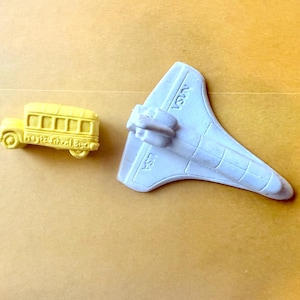 May include: A yellow school bus and a light gray space shuttle toy are displayed on a light brown surface. The space shuttle has the letters "NASA" printed on it. The school bus has details like windows and wheels.