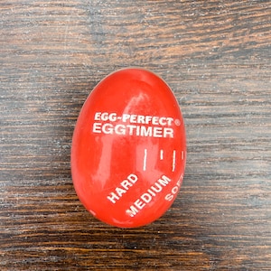 May include: A red egg-shaped kitchen timer with the text "EGG-PERFECT EGGTIMER". The timer has markings for "HARD", "MEDIUM", and "SOFT". The timer is designed to help cook eggs to the desired doneness.