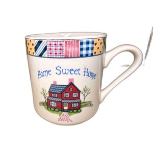May include: Cream-colored ceramic mug with a curved handle. The mug features a colorful patchwork design around the rim and a charming illustration of a red house with a blue roof, trees, and the words "Home Sweet Home."