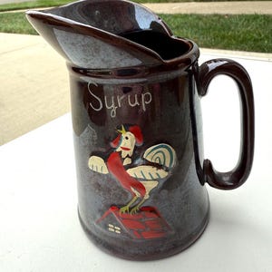 May include: Brown ceramic syrup pitcher with a handle and spout. The pitcher features a rooster design with the word "Syrup" in white script. The rooster is red, white, and blue, standing on a red roof.