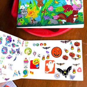 May include: Open sticker book with various stickers, including Halloween-themed designs like jack-o'-lanterns, bats, and candy corn. The book also features colorful, glittery stickers and a cover with woodland creatures and flowers.