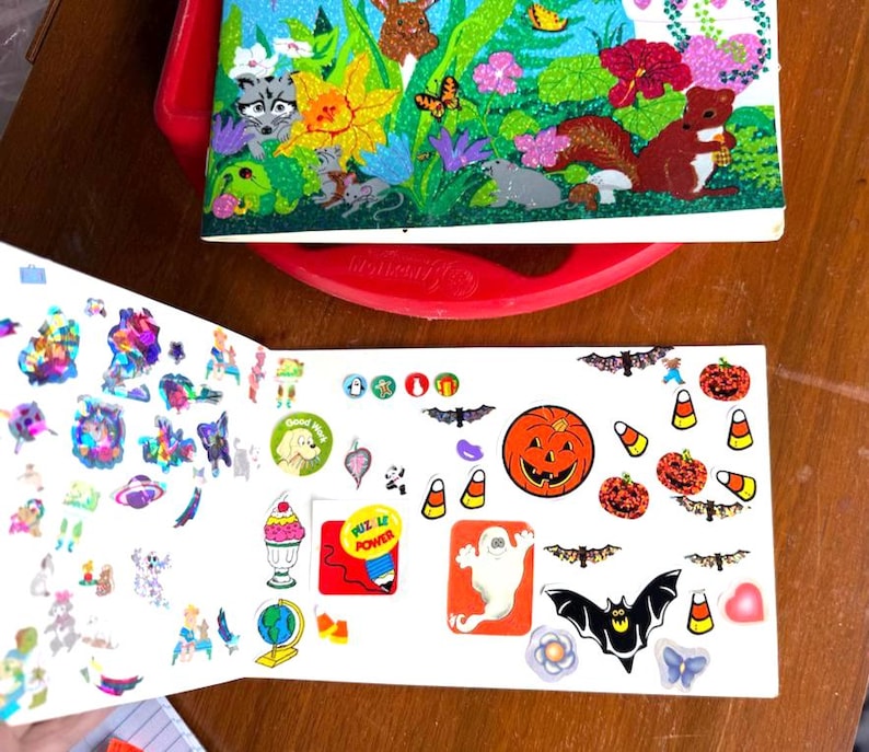 May include: Open sticker book with various stickers, including Halloween-themed designs like jack-o'-lanterns, bats, and candy corn. The book also features colorful, glittery stickers and a cover with woodland creatures and flowers.
