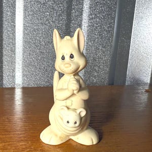 May include: A cream-colored kangaroo figurine with a joey in its pouch. The kangaroo has its hands clasped together and a smiling expression. The figurine is made of a smooth material and has a glossy finish.
