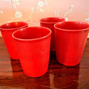 May include: A set of four solid red tumblers with a smooth, matte finish. Each tumbler features a simple, cylindrical shape with a slightly tapered base and an open top. The cups are arranged in a cluster, displaying a consistent bright red color.