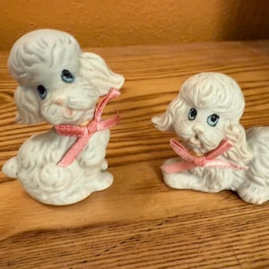 May include: Two white ceramic poodle figurines with blue eyes and pink bows around their necks. One poodle is sitting upright, while the other is lying down. The figurines are on a wooden surface.