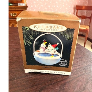 May include: A Keepsake Ornament box featuring a Peanuts-themed holiday scene with Snoopy and Woodstock snow figures. The box is brown and gold with the words "Keepsake Ornament" and "Magic Collector's Series".