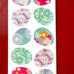 May include: A sheet of colorful Easter egg stickers. Each egg features a unique design, including floral patterns, paisley, and a bird. The eggs are in shades of pink, green, purple, and blue, with glitter accents. The background is white, and the sheet is on a red surface.