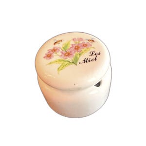 May include: A white ceramic jar with a lid featuring a floral design and the text "Les Miel". The lid has pink flowers, green leaves, and two bees. The jar is round with a small opening.