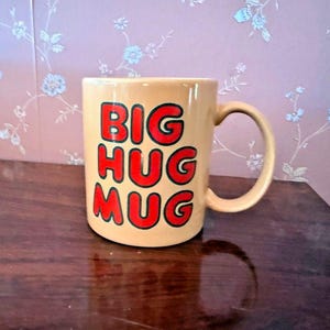 May include: A light tan ceramic mug with a rounded handle, featuring the text BIG HUG MUG printed in bold red letters with a black outline on the front. The mug has a smooth, glossy finish and a cylindrical shape with a flat base.