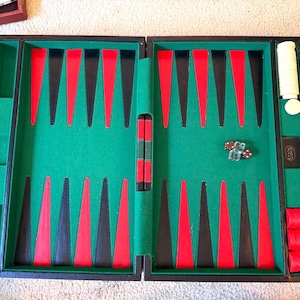 May include: A backgammon board game set featuring a green felt interior with alternating red and black triangular points. The set includes white and red circular checkers, two translucent dice, and a black doubling cube with white text.