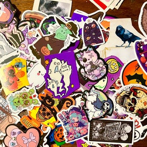 May include: A collection of colorful Halloween themed stickers featuring various characters, including ghosts, cats, pumpkins, and a skull. The stickers are arranged in a random pattern on a wooden surface.