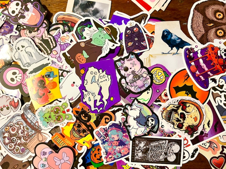May include: A collection of colorful Halloween themed stickers featuring various characters, including ghosts, cats, pumpkins, and a skull. The stickers are arranged in a random pattern on a wooden surface.