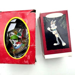 May include: Two Christmas ornaments in red boxes. One features Marvin the Martian in a sleigh, the other Bugs Bunny. The Bugs Bunny ornament is gray with a white hat and gloves. Both boxes have a cartoon theme.