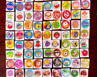 May include: A sheet of colorful stickers with various cartoon characters and phrases. The stickers are round and feature a variety of themes, including animals, food, and holidays. Some of the phrases on the stickers include "Go for it!" "Stick to it!" "Super Star!" and "Berry Good!"