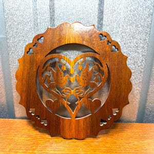 May include: A round, wooden decorative piece with a heart-shaped cutout in the center. The heart design features two birds facing each other, with a bow at the bottom. The outer edge has a scalloped design with heart-shaped cutouts. The wood has a warm, brown tone.