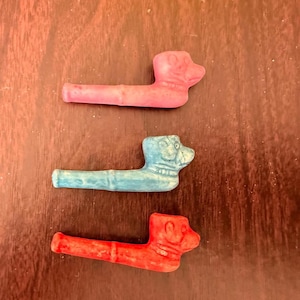May include: Three small, dog-shaped pipes in pink, blue, and red are arranged on a dark wood surface. Each pipe features a detailed dog head design and a long, cylindrical stem. The pipes are likely decorative or novelty items.