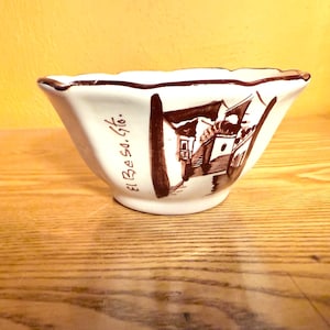 May include: White ceramic bowl with a scalloped rim and a brown illustration of buildings. The bowl has the text "El Beso. Gto." in brown. The bowl is on a wooden surface.