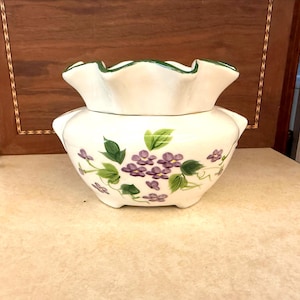 May include: A white ceramic container with a ruffled rim and a lid. The container is decorated with purple flower and green leaf designs. The rim is trimmed with green paint.