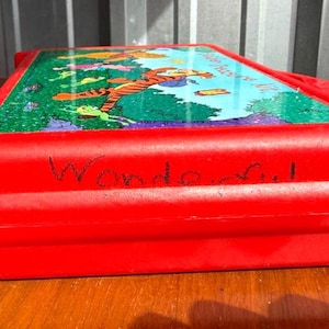 May include: A red plastic lunchbox featuring a colorful illustration of Tigger from Winnie the Pooh. The lunchbox has the word "Wonderful" written on the side. The top of the lunchbox has a handle.