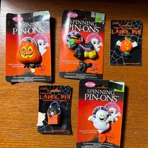 May include: A collection of Halloween-themed lapel pins and spinning pin-ons. The pins feature a jack-o'-lantern, a witch on a broom, a ghost, and a black cat. The pins are on orange and black cards.