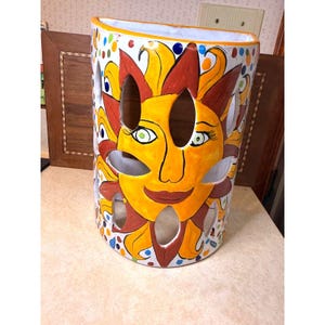May include: A cylindrical ceramic vase with a smiling sun face design. The vase is painted with yellow, orange, and red colors, with cut-out shapes for light. The vase is decorated with colorful polka dots.