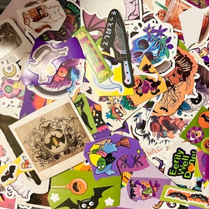 May include: A collection of colorful Halloween themed stickers featuring various designs, including bats, ghosts, pumpkins, and witches. The stickers are arranged in a random pattern on a white surface.