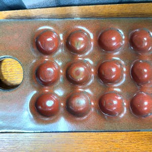 May include: A rectangular, ceramic escargot plate with 12 indentations for snails. The plate is a mottled reddish-brown color with a circular handle on one end. The indentations are a darker red.