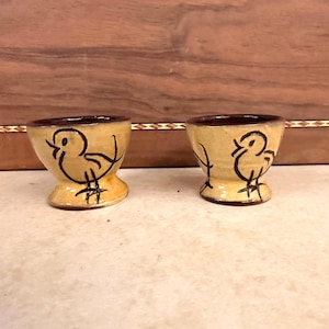 May include: Two small, beige ceramic egg cups with a flared base and a dark brown interior rim. Each cup features a hand-painted, black line drawing of a stylized bird on the exterior. The cups have a smooth, glossy finish and a rounded shape.
