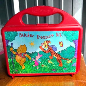 May include: Red plastic "Sticker Treasure Kit" lunchbox featuring Winnie the Pooh, Tigger, and Piglet. The lunchbox has a handle and a colorful illustration of the characters in a forest scene. The text "Sticker Treasure Kit" is displayed on the front.