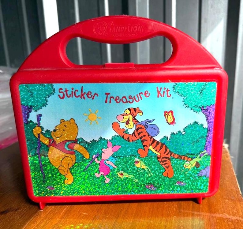 May include: Red plastic "Sticker Treasure Kit" lunchbox featuring Winnie the Pooh, Tigger, and Piglet. The lunchbox has a handle and a colorful illustration of the characters in a forest scene. The text "Sticker Treasure Kit" is displayed on the front.