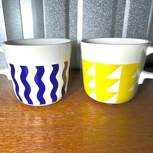 May include: Two white ceramic mugs with curved handles. One mug features a blue wavy pattern, and the other has a yellow geometric design. Both mugs are on a wooden surface.