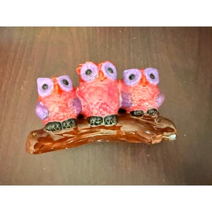 May include: A ceramic figurine featuring three owls perched on a brown branch. The owls have red bodies, purple wings, and large purple eyes. The branch is a dark brown color, and the figurine has a glossy finish.