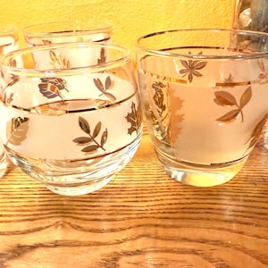 May include: Set of clear, round drinking glasses with a frosted band and gold leaf design. The glasses have a gold rim and are displayed on a wooden surface. The glassware is suitable for serving beverages.