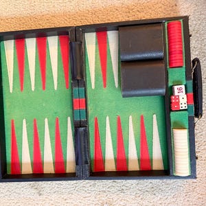 May include: A black backgammon board game set featuring a green felt interior with red and white triangular points. The set includes two black dice cups, a stack of red checkers, a stack of white checkers, and two dice with the number 16 visible.