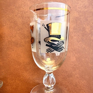 May include: Clear glass goblet with a gold rim and a decorative stem. The glass features frosted panels with black and gold illustrations of a top hat, gloves, and other vintage accessories. A classic barware item.