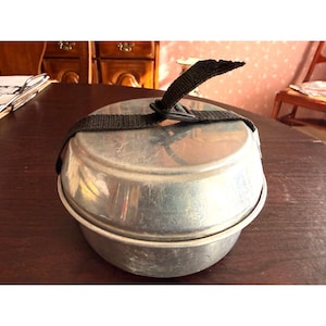May include: A small, round, aluminum camping mess kit with a black strap and buckle. The kit is closed and appears to be used for carrying food. The metal has a slightly worn appearance.