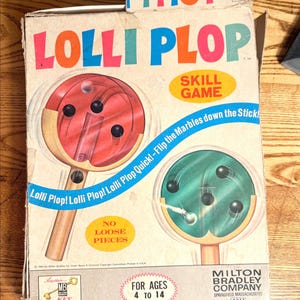May include: Vintage "Lolli Plop" skill game box. The box features the game's name in colorful letters, illustrations of the game, and text that reads "Flip the Marbles down the Stick!" and "For Ages 4 to 14."