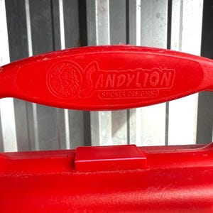 May include: Close-up of a red plastic handle with the embossed text "SANDYLION (STICKER DESIGNS)" and a lion logo. The handle has a large oval opening and a rectangular tab. The background is a corrugated metal surface.