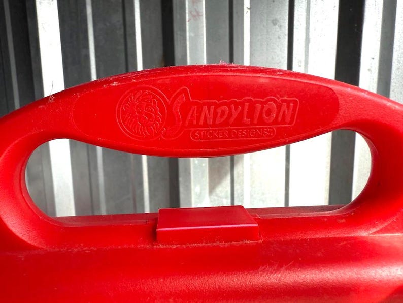 May include: Close-up of a red plastic handle with the embossed text "SANDYLION (STICKER DESIGNS)" and a lion logo. The handle has a large oval opening and a rectangular tab. The background is a corrugated metal surface.