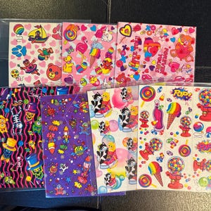May include: A collection of colorful sticker sheets featuring various designs. The stickers include images of bears, cats, rockets, and other whimsical illustrations. The color palette is vibrant, with pinks, purples, and blues dominating the designs. Some sheets are in clear plastic sleeves.