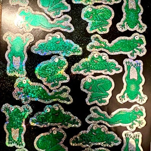 May include: A sheet of green frog stickers with a holographic finish. The frogs are in various poses, including sitting, jumping, and swimming.
