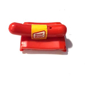 May include: A red plastic toy whistle shaped like an Oscar Mayer hot dog. The whistle has a yellow label with the words "Oscar Mayer" printed on it.