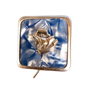 May include: A square pin with a silver frame and a blue, textured background. A gold-colored ship is in the center. The pin has a straight pin backing.