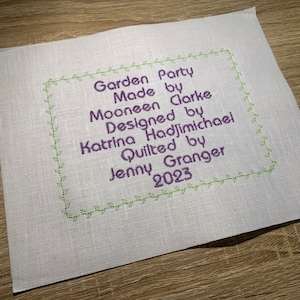 May include: A white linen fabric with a purple and green embroidered label that reads "Garden Party Made by Mooneen Clarke Designed by Katrina Hadjimichael Quilted by Jenny Granger 2023".