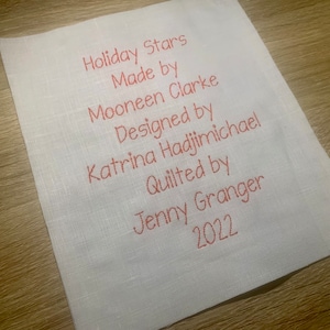 May include: A white fabric label with red text that reads "Holiday Stars Made by Mooneen Clarke Designed by Katrina Hadjimichael Quilted by Jenny Granger 2022".