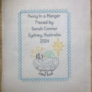 May include: A white linen fabric with a blue and green embroidered border. The centre of the fabric features a yellow star and an embroidered image of a baby in a manger. The text "Away in a Manger" is embroidered in grey, along with "Pieced by Sarah Connor, Sydney, Australia 2024".