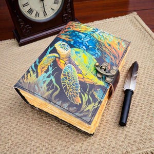 May include: A leather journal with a sea turtle design. The cover displays a colourful illustration of a turtle amongst coral and seaweed. A decorative clasp and a feather pen are also visible.