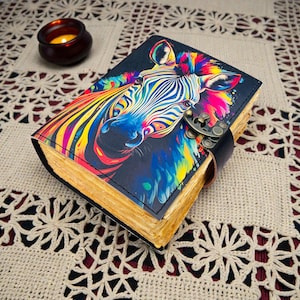 May include: A leather-bound journal with a vibrant, colorful zebra illustration on the cover. The zebra's stripes are rendered in a rainbow of hues. The journal has a black leather strap closure and aged paper pages.