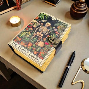 May include: A rectangular journal with a black cover featuring a skeleton surrounded by various potted plants and flowers. The cover displays the text THE PLANT LOVER and a Roman numeral XI. It includes a brass-colored metal latch and yellow pages.