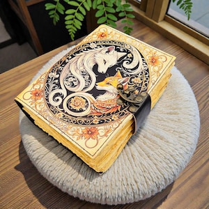May include: A rectangular journal with a cream-colored cover featuring a central oval illustration of a white fox and an orange fox surrounded by floral patterns. The book is secured with a dark metal latch and has thick, aged-looking pages.